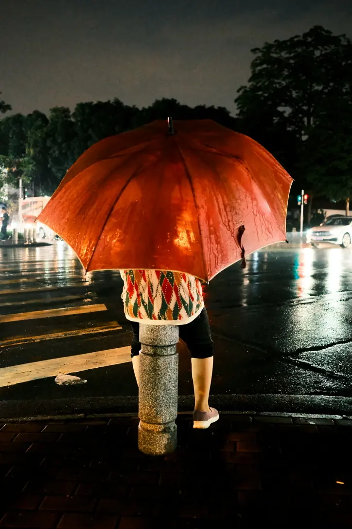 Una persona de espaldas bajo un paraguas rojo, junto a un paso de cebra mojado, de noche
